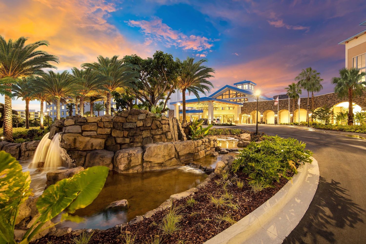 Twilight view of Loews Sapphire Falls Resort entrance with illuminated palm trees and waterfall at a favorite Universal Studio hotels in Orlando, Florida