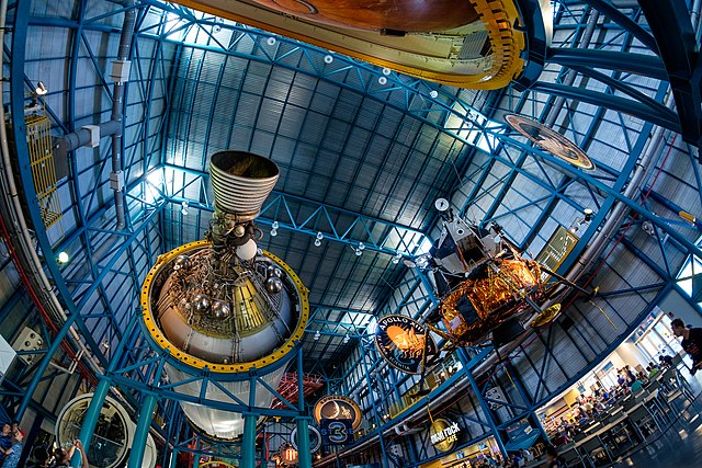 A wide-angle view of the interior of the Kennedy Space Center, featuring large rocket engines and spacecraft exhibits suspended from the high ceiling.