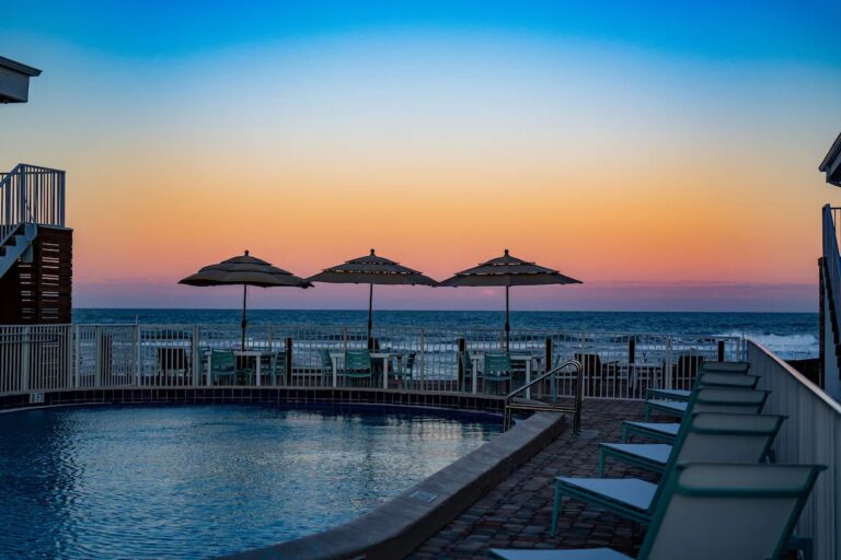 A beautiful sunset view from the poolside at Melbourne Beach Florida Resort, featuring umbrellas, lounge chairs, and the ocean in the background.