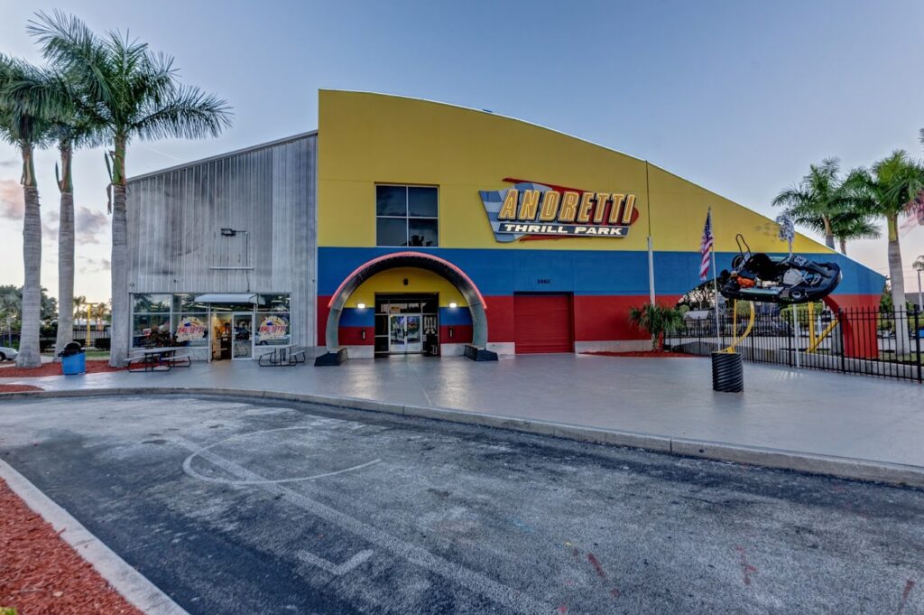 Exterior of Andretti Thrill Park with a yellow and blue building, a colorful entrance arch, and palm trees, featuring a race car sculpture and clear sky.