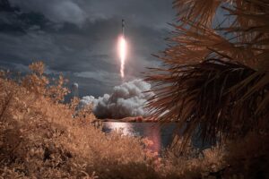 An infrared view of a rocket launching amid clouds, framed by palm leaves and reflected in a body of water, a popular space coast attraction.