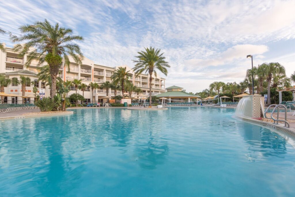 View of a resort pool with clear blue water, surrounded by palm trees and lounge chairs, located at Holiday Inn Cape Canaveral in Florida. A bright, sunny sky reflects on the peaceful setting.