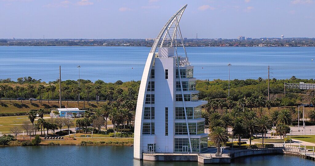 A seven-story landmark building, Exploration Tower, located in Port Canaveral, with a distinctive sail-like design, surrounded by greenery and water, popular of the many space coast attractions.