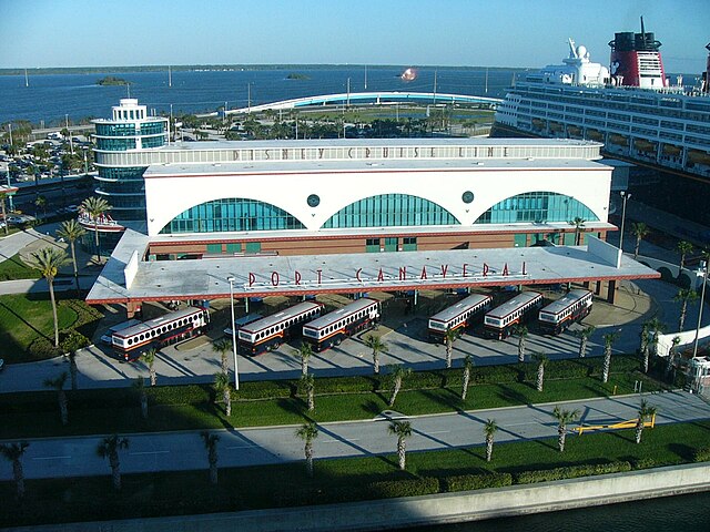 Aerial view of Port Canaveral showcasing the terminal building and adjacent parked shuttles, with a cruise ship docked in the background.