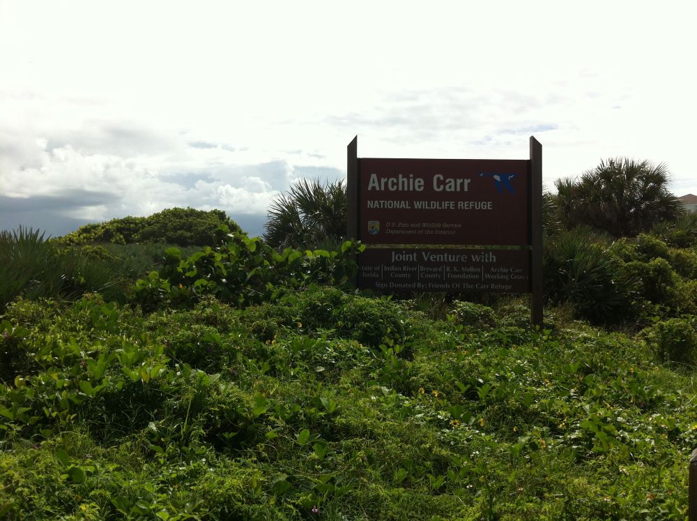 A sign for the Archie Carr National Wildlife Refuge, surrounded by greenery and under a cloudy sky.