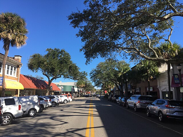 A wide street in historic downtown Melbourne, lined with parked cars on both sides and trees providing shade, under a clear blue sky.