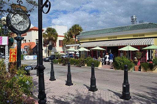 A street view of Cocoa Village with decorative streetlamps, a colorful building with outdoor seating, palm trees, and a visible clock.