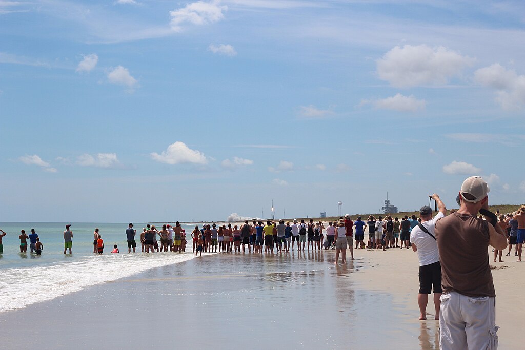 A large crowd gathered at Playalinda Beach, standing along the shoreline with waves lapping at their feet, under a partly cloudy sky.